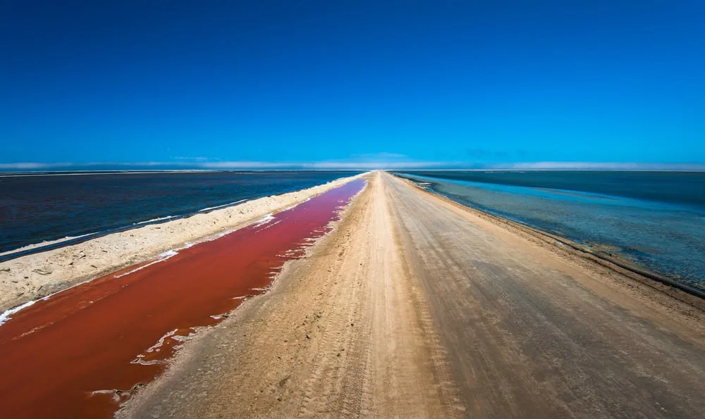 Estrada entre salina com salmoura avermelhada e água azul